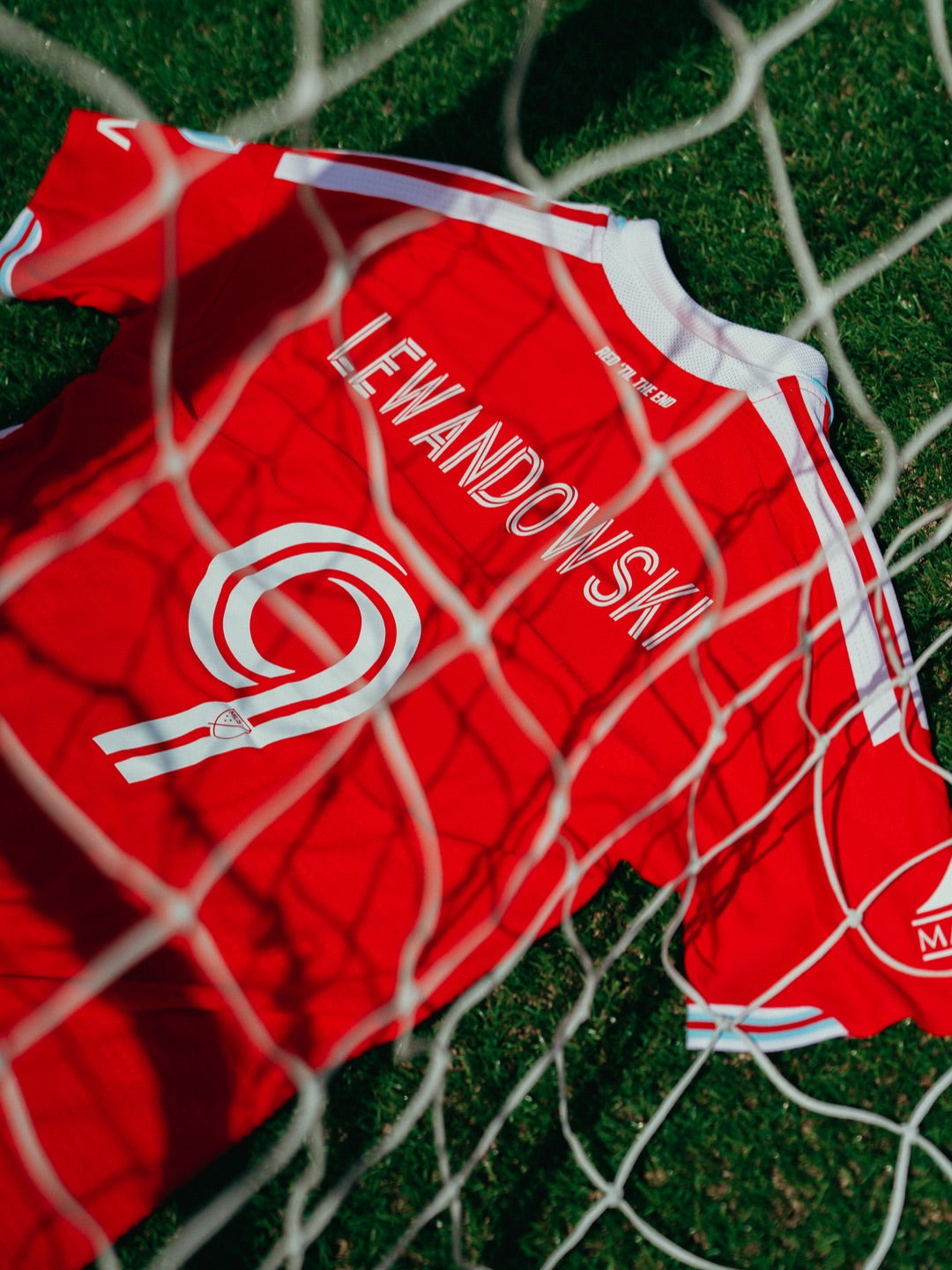 A red soccer jersey with 'LEWANDOWSKI' and a large number 9 printed in white, laid on green grass behind a white soccer net.